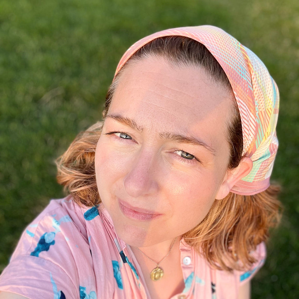 Woman wearing a colorful headscarf and pink top with blue patterns against a green grass background